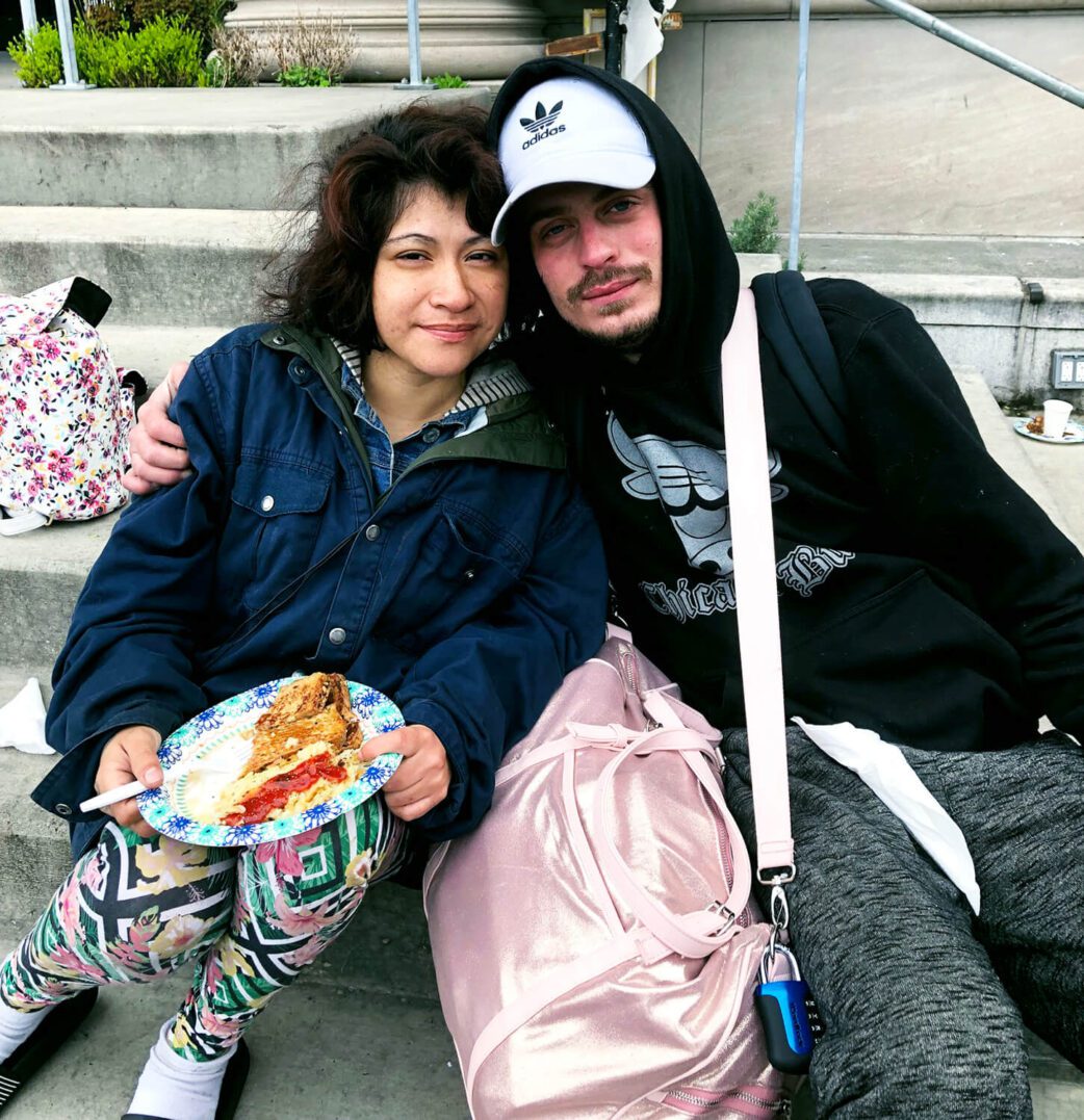 Two friends sitting closely with food and backpacks outdoors.